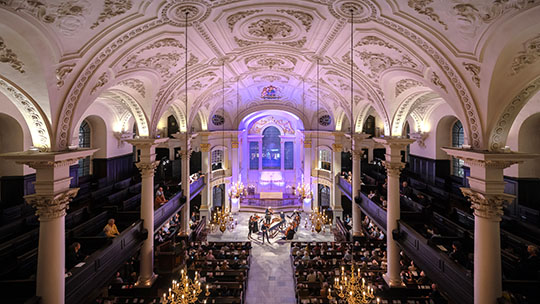 Inside St Martins-in-the-fields from high up. White, patterned ceiling with columns. Audience in pews watching string ensemble