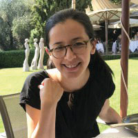 Louisa Price Headshot. White woman with dark hair pulled back and glasses. Wearing a black shirt. Sitting dow.