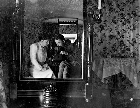 Black and white self-portrait. Mirror reflection of 2 people, floral background in room.